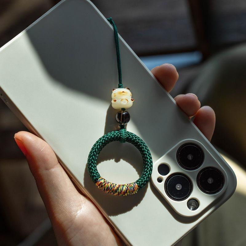 Dark green Y290 lucky cat handwoven ring strap hanging from a smartphone in sunlight